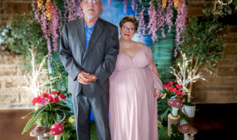 A man in a gray suit stands beside a woman with an intellectual disability (IDD) in a soft pink gown at Home Not Alone's prom event hosted by Milestone at Laube Hall, Pennsylvania, posing in front of a floral backdrop with colorful flowers and greenery.