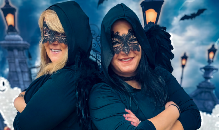 Two individuals in gothic black costumes with feathered capes and lace masks pose back-to-back, embracing the Halloween spirit. Home Not Alone Halloween event 2024.