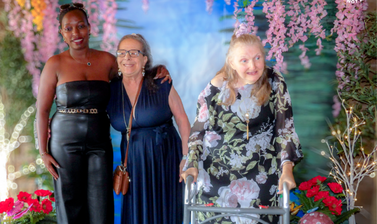 Three women, including two individuals with intellectual disabilities (IDD) and one caregiver, stand together celebrating at the Home Not Alone prom hosted by Milestone at Laube Hall, Pennsylvania, against a colorful, nature-themed backdrop.