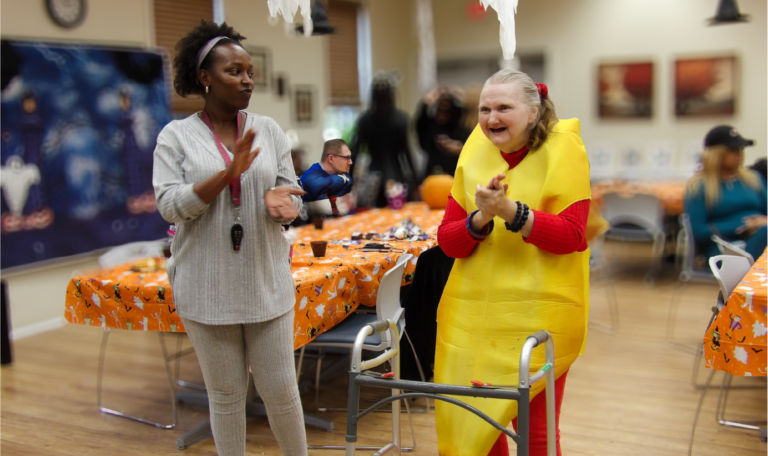 A caregiver and a client, with one dressed as a banana, share a happy moment clapping together, adding to the festive energy. Home Not Alone Halloween event 2024.