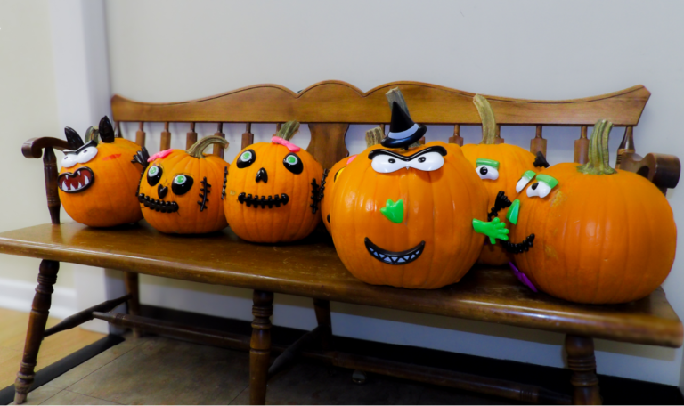 A row of carved pumpkins with expressive, humorous faces sits on a wooden bench, each decorated with unique features like hats and faces, adding a playful touch to the event. Home Not Alone Halloween event 2024.