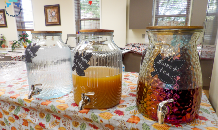 A drink station featuring three glass dispensers filled with water, apple cider, and sweet tea, labeled with charming leaf-shaped signs on a fall-themed table at the Thanksgiving 2024 'Friendsgiving' Event by Home Not Alone Caregiver Services for individuals with intellectual disability (IDD) within residential group homes and accessible family group homes in Allegheny County, Pittsburgh, Pennsylvania.