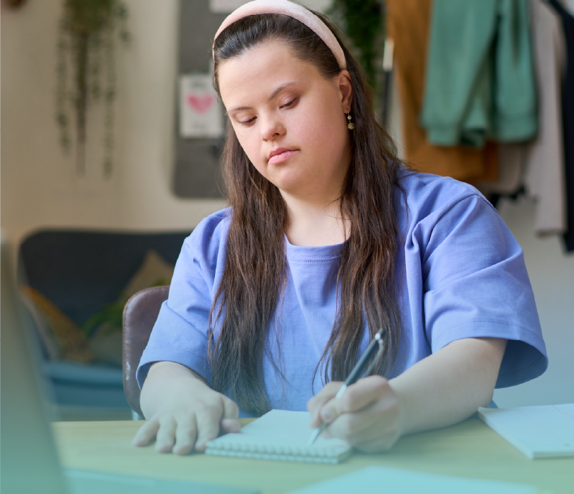 A young woman with Down syndrome writing in a notebook at a wooden desk, deeply focused. The setting includes a cozy room with decorations and a warm ambiance, fostering creativity and independence. Provided by Home Not Alone Caregiver Services for individuals with intellectual disability (IDD) within Community for Disabled Adults and accessible family group homes in Allegheny County, Pittsburgh, Pennsylvania.