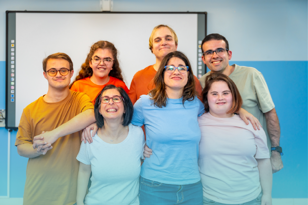 A group of seven individuals, including men and women with intellectual and developmental disabilities, smile together in a classroom. They stand closely, showing a sense of friendship and support. This image highlights inclusion during Developmental Disabilities Awareness Month (DDAM). Provided by Home Not Alone Caregiver Services for individuals with intellectual disability (IDD) within Community for Disabled Adults and accessible family group homes in Allegheny County, Pittsburgh, Pennsylvania.