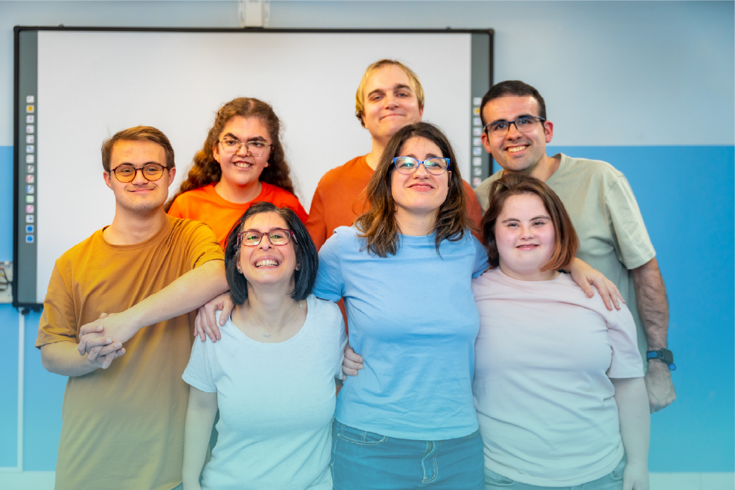 A group of seven individuals, including men and women with intellectual and developmental disabilities, smile together in a classroom. They stand closely, showing a sense of friendship and support. This image highlights inclusion during Developmental Disabilities Awareness Month (DDAM). Provided by Home Not Alone Caregiver Services for individuals with intellectual disability (IDD) within Community for Disabled Adults and accessible family group homes in Allegheny County, Pittsburgh, Pennsylvania.