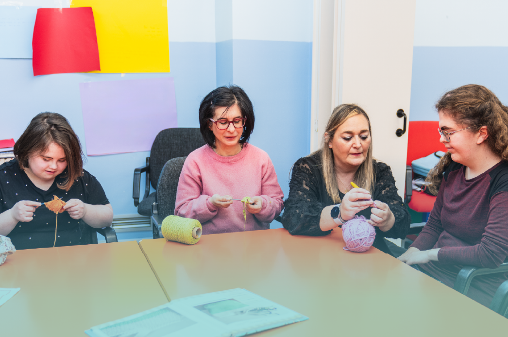 Four women, including individuals with intellectual and developmental disabilities, sit around a table in a classroom, knitting with colorful yarn. One appears to be a DSP/Caregiver teaching a disabled women. This image represents creativity and empowerment during Developmental Disabilities Awareness Month (DDAM). Provided by Home Not Alone Caregiver Services for individuals with intellectual disability (IDD) within Community for Disabled Adults and accessible family group homes in Allegheny County, Pittsburgh, Pennsylvania.