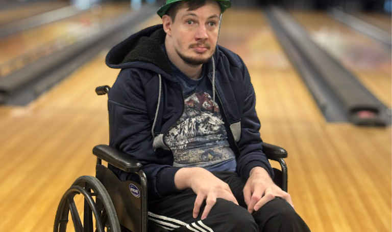 A man in a green glittery St. Patrick's Day hat sits in a wheelchair on a bowling lane, looking ahead with a calm expression. He wears a navy jacket and striped sweatpants. Provided by Home Not Alone Caregiver Services for individuals with intellectual disability (IDD) within Community for Disabled Adults and accessible family group homes in Allegheny County, Pittsburgh, Pennsylvania.