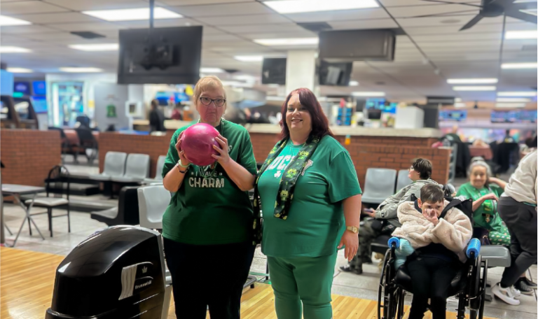Two women stand smiling at a bowling lane, one holding a pink bowling ball. Both are dressed in green for St. Patrick’s Day. Behind them, a young woman in a wheelchair and others in green sit and watch. Provided by Home Not Alone Caregiver Services for individuals with intellectual disability (IDD) within Community for Disabled Adults and accessible family group homes in Allegheny County, Pittsburgh, Pennsylvania.