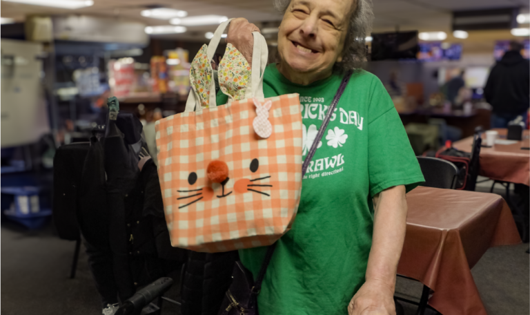 A cheerful woman in a green St. Patrick’s Day shirt holds a pink gingham bag decorated like a bunny face. She stands beside a walker and smiles warmly in a social setting filled with tables and chairs. Provided by Home Not Alone Caregiver Services for individuals with intellectual disability (IDD) within Community for Disabled Adults and accessible family group homes in Allegheny County, Pittsburgh, Pennsylvania.