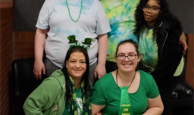 Five women dressed in green and tie-dye outfits gather for a group photo on St. Patrick’s Day, smiling brightly. They pose against a brick wall in a bowling alley. Provided by Home Not Alone Caregiver Services for individuals with intellectual disability (IDD) within Community for Disabled Adults and accessible family group homes in Allegheny County, Pittsburgh, Pennsylvania.