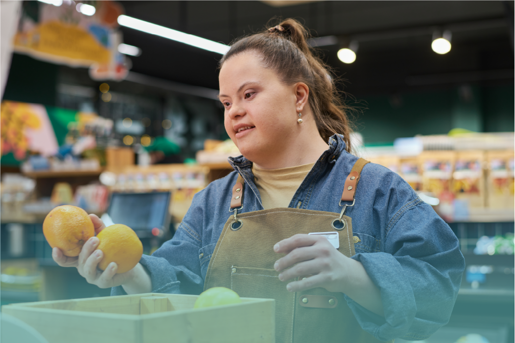 A young woman with Down syndrome works in a grocery store, wearing a denim jacket and a brown apron. She is sorting oranges with a focused expression. The background shows shelves stocked with various products. This image highlights workplace inclusion and independence for individuals with Down syndrome on World Down Syndrome Day. Provided by Home Not Alone Caregiver Services for individuals with intellectual disability (IDD) within Community for Disabled Adults and accessible family group homes in Allegheny County, Pittsburgh, Pennsylvania.