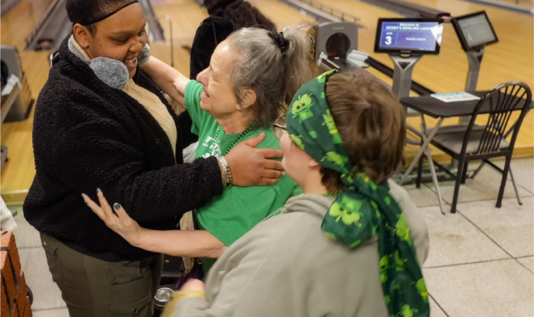 Three women share a joyful group hug near the bowling lanes. One wears earmuffs, another a green shirt, and a third has a shamrock-themed scarf. The background features bowling lanes and colorful screens. Provided by Home Not Alone Caregiver Services for individuals with intellectual disability (IDD) within Community for Disabled Adults and accessible family group homes in Allegheny County, Pittsburgh, Pennsylvania.