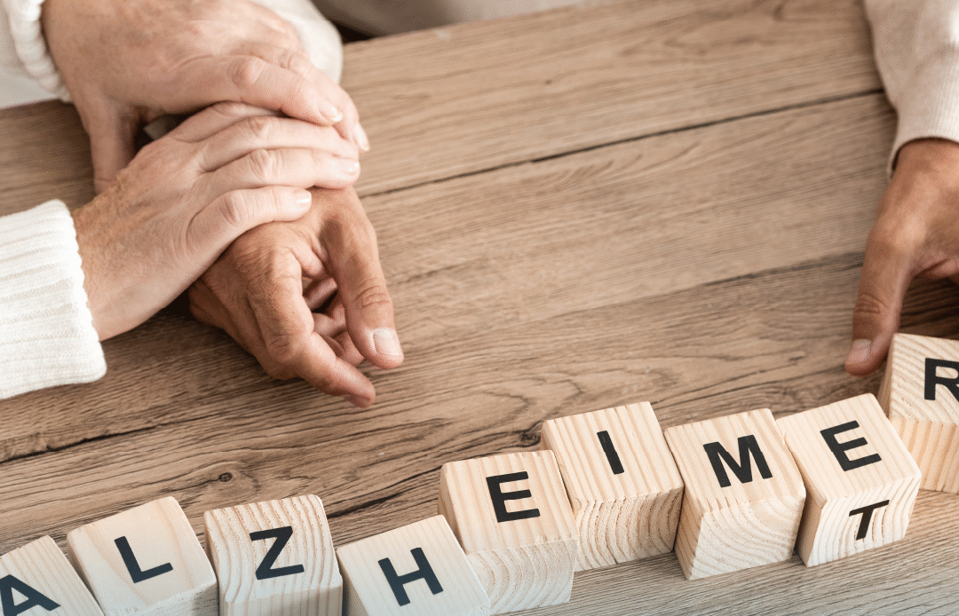 A close-up of two people gently holding hands on a wooden table, next to wooden letter blocks spelling “ALZHEIMER,” representing caregiving and emotional support. Provided by Home Not Alone Caregiver Services for individuals with intellectual disability (IDD) within Community for Disabled Adults and accessible family group homes in Allegheny County, Pittsburgh, Pennsylvania.