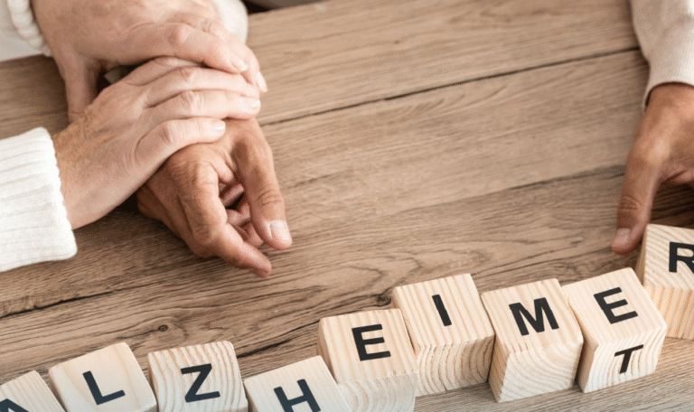 A close-up of two people gently holding hands on a wooden table, next to wooden letter blocks spelling “ALZHEIMER,” representing caregiving and emotional support. Provided by Home Not Alone Caregiver Services for individuals with intellectual disability (IDD) within Community for Disabled Adults and accessible family group homes in Allegheny County, Pittsburgh, Pennsylvania.