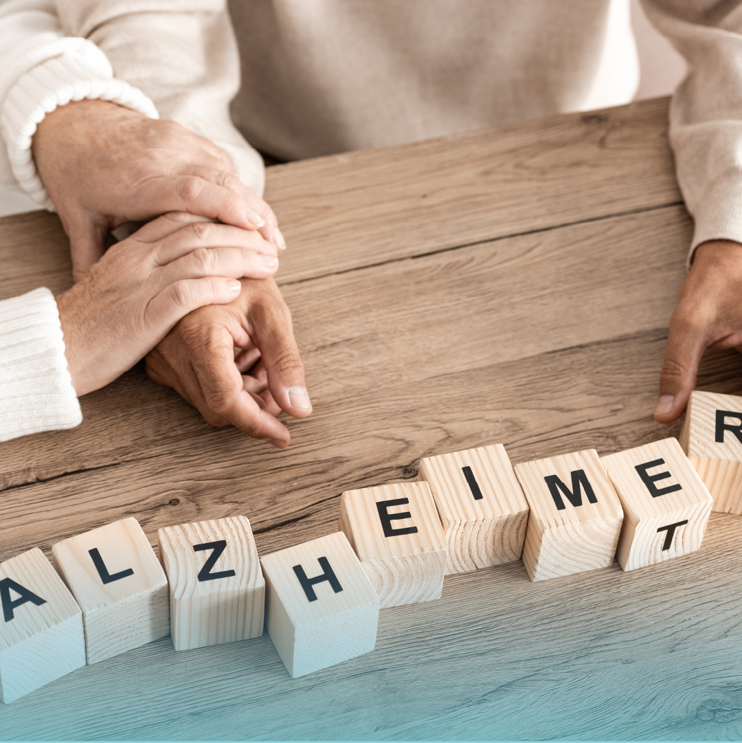 A close-up of two people gently holding hands on a wooden table, next to wooden letter blocks spelling “ALZHEIMER,” representing caregiving and emotional support. Provided by Home Not Alone Caregiver Services for individuals with intellectual disability (IDD) within Community for Disabled Adults and accessible family group homes in Allegheny County, Pittsburgh, Pennsylvania.
