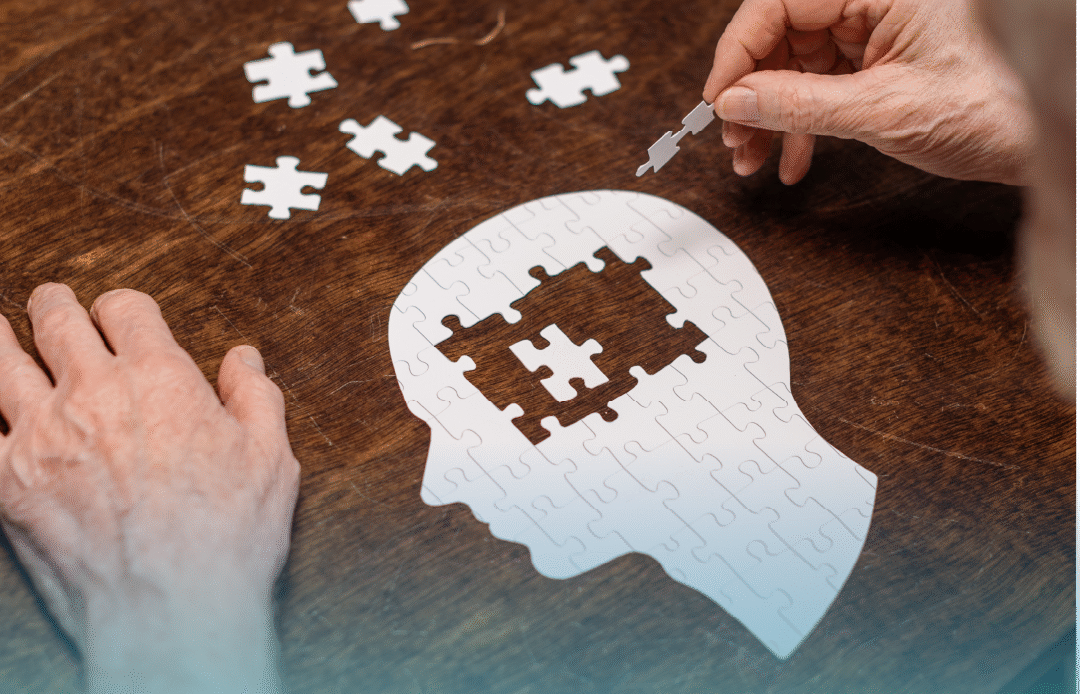 A puzzle in the shape of a human head on a wooden table, with hands placing the final piece inside the brain area, symbolizing memory loss and Alzheimer’s. Provided by Home Not Alone Caregiver Services for individuals with intellectual disability (IDD) within Community for Disabled Adults and accessible family group homes in Allegheny County, Pittsburgh, Pennsylvania.