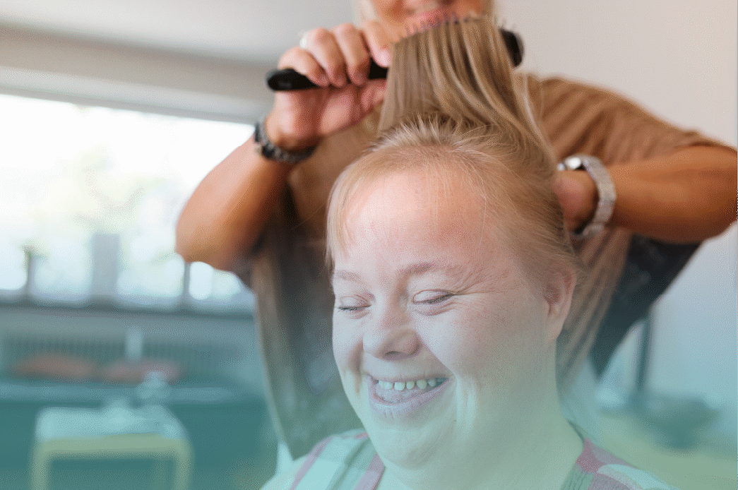 A woman with an intellectual disability smiles joyfully as a caregiver brushes her hair, capturing a warm moment of personal grooming and confidence. Provided by Home Not Alone Caregiver Services for individuals with intellectual disability and Autism (ID/A) within Community for Disabled Adults and accessible family group homes in Allegheny County, Pittsburgh, Pennsylvania.
