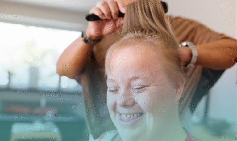 A woman with an intellectual disability smiles joyfully as a caregiver brushes her hair, capturing a warm moment of personal grooming and confidence. Provided by Home Not Alone Caregiver Services for individuals with intellectual disability and Autism (ID/A) within Community for Disabled Adults and accessible family group homes in Allegheny County, Pittsburgh, Pennsylvania.