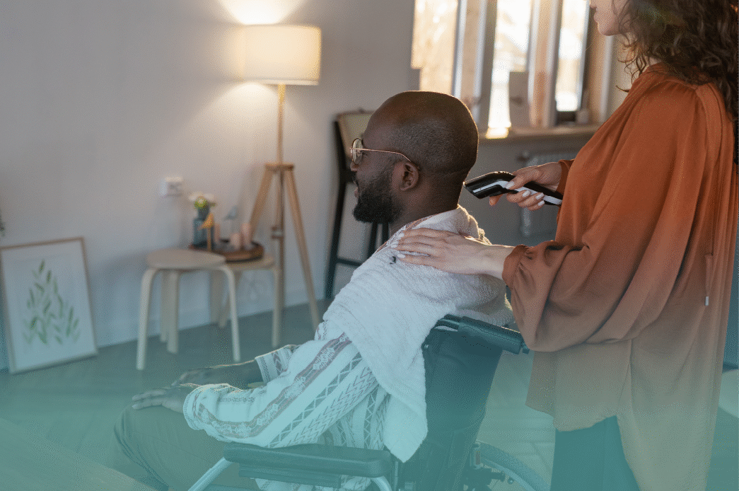 A caregiver gently places a towel around a man in a wheelchair and prepares to groom him with electric clippers, providing supportive personal care in a calm home environment. Provided by Home Not Alone Caregiver Services for individuals with intellectual disability and Autism (ID/A) within Community for Disabled Adults and accessible family group homes in Allegheny County, Pittsburgh, Pennsylvania.
