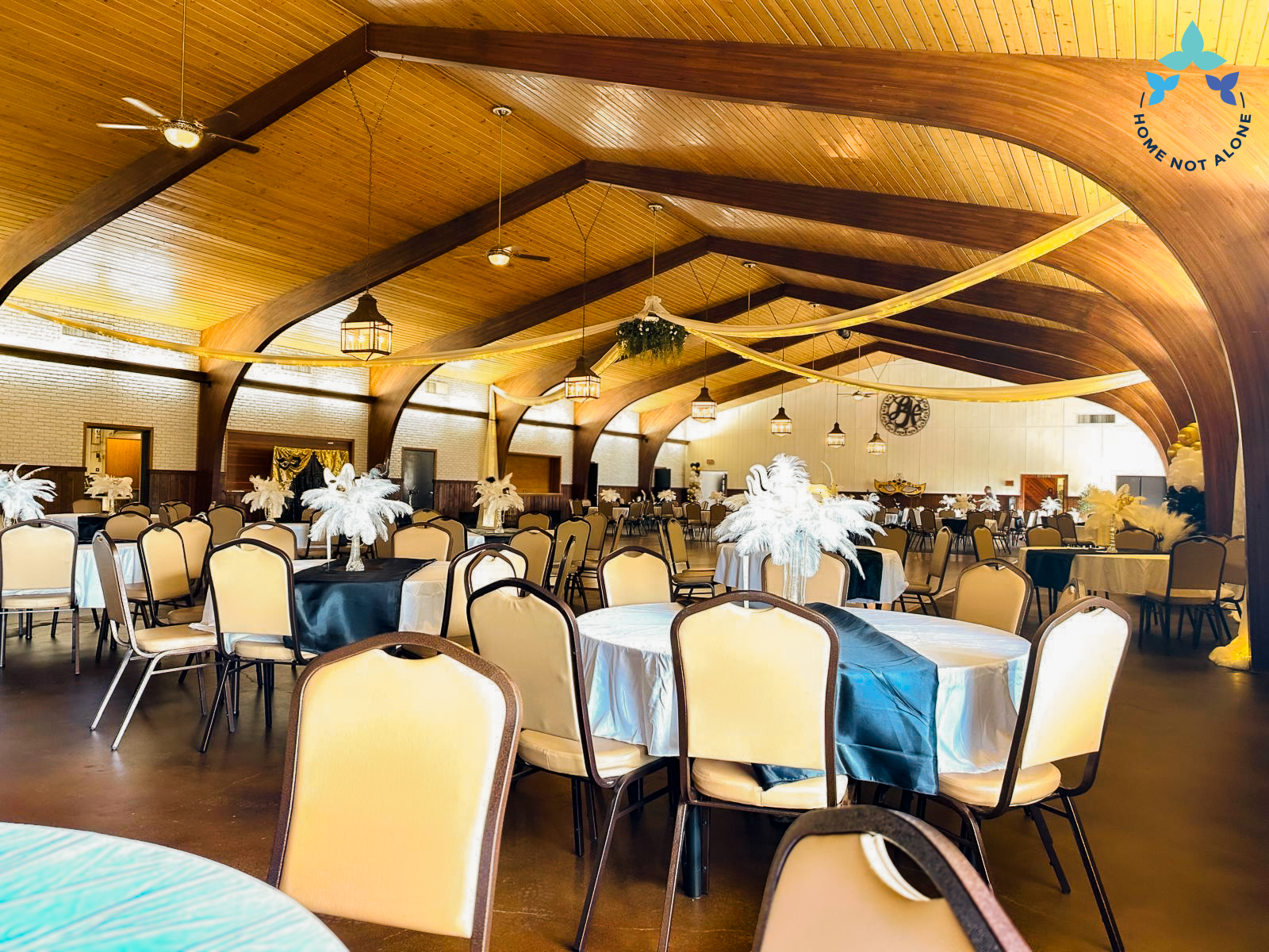 A wide view of the ballroom filled with decorated tables, black table runners, and white feather centerpieces prepared for the prom event. Provided by Home Not Alone Caregiver Services for individuals with intellectual disability and Autism (ID/A) within Community for Disabled Adults and accessible family group homes in Allegheny County, Pittsburgh, Pennsylvania.