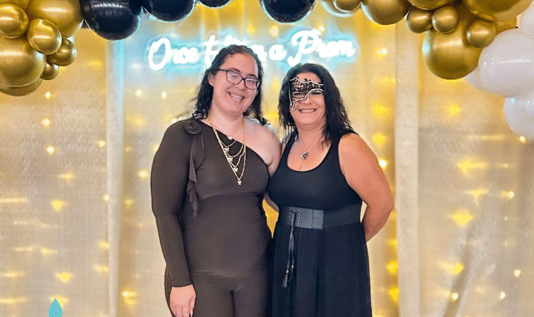 Two women standing under a black and gold balloon arch at the prom, one in a brown jumpsuit and the other in a black dress with a masquerade mask, smiling for a photo. Provided by Home Not Alone Caregiver Services for individuals with intellectual disability and Autism (ID/A) within Community for Disabled Adults and accessible family group homes in Allegheny County, Pittsburgh, Pennsylvania.