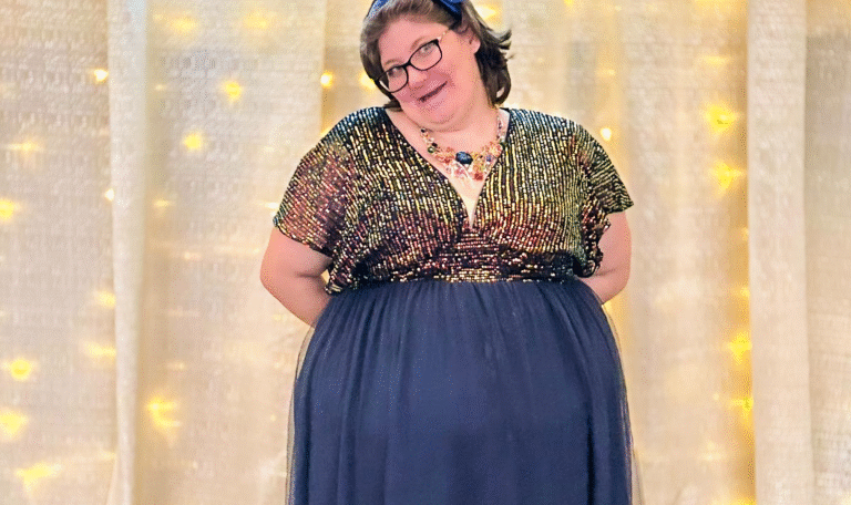 A woman standing under the prom backdrop in a navy gown with a glittering top and colorful necklace, smiling brightly in front of the golden curtain and “Once Upon a Prom” sign. Provided by Home Not Alone Caregiver Services for individuals with intellectual disability and Autism (ID/A) within Community for Disabled Adults and accessible family group homes in Allegheny County, Pittsburgh, Pennsylvania.