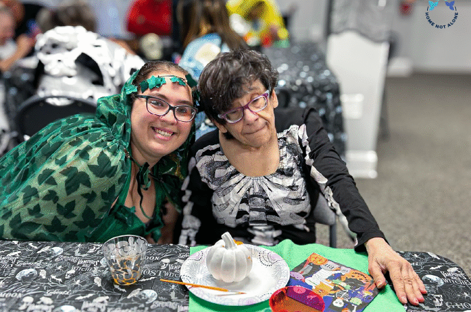 Two women enjoying a lighthearted moment during the Home Not Alone Halloween celebration, smiling and sharing joy in a welcoming environment. Provided by Home Not Alone Caregiver Services for individuals with intellectual disability and Autism (ID/A) within Community for Disabled Adults and accessible family group homes in Allegheny County, Pittsburgh, Pennsylvania.