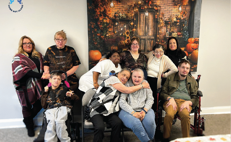 A group photo of caregivers and individuals from Home Not Alone gathered in front of a cozy fall backdrop, sharing warm smiles at the Thanksgiving celebration. Provided by Home Not Alone Caregiver Services for individuals with intellectual disability and Autism (ID/A) within Community for Disabled Adults and accessible family group homes in Allegheny County, Pittsburgh, Pennsylvania.