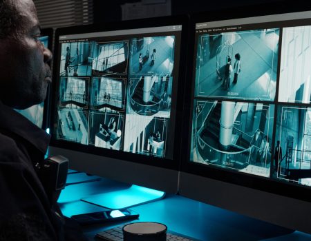A man attentively monitoring multiple security camera feeds on dual computer screens, reflecting real-time observation for home safety. Provided by Home Not Alone Caregiver Services for individuals with intellectual disability and Autism (ID/A) within Community for Disabled Adults and accessible family group homes in Allegheny County, Pittsburgh, Pennsylvania.
